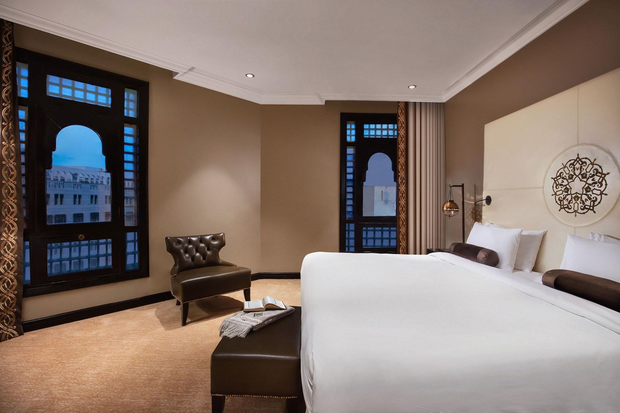Sofitel Shahd Al Madinah Hotel Medina Buitenkant foto The photo shows a modern hotel room featuring a large bed with white linens, a dark upholstered headboard, and a tufted armchair in the corner. There are large windows with decorative frames that allow natural light to enter the room, and the walls a