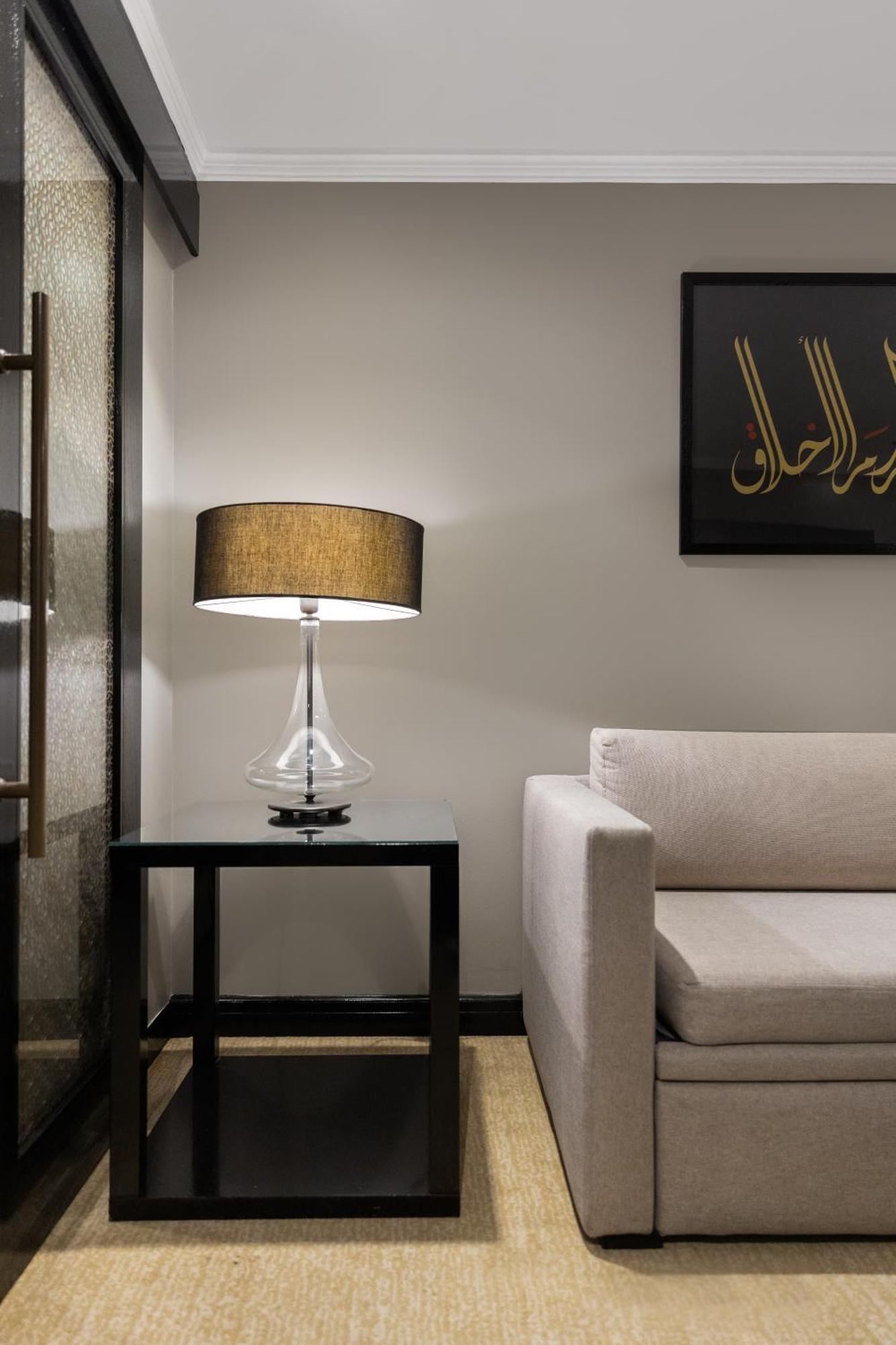 Sofitel Shahd Al Madinah Hotel Medina Buitenkant foto The photo depicts a modern interior setting. On the left, there is a glass table lamp with a cylindrical body and a round, dark-colored lampshade. The lamp is placed on a sleek black side table. To the right, there’s a light-colored sofa with a minim
