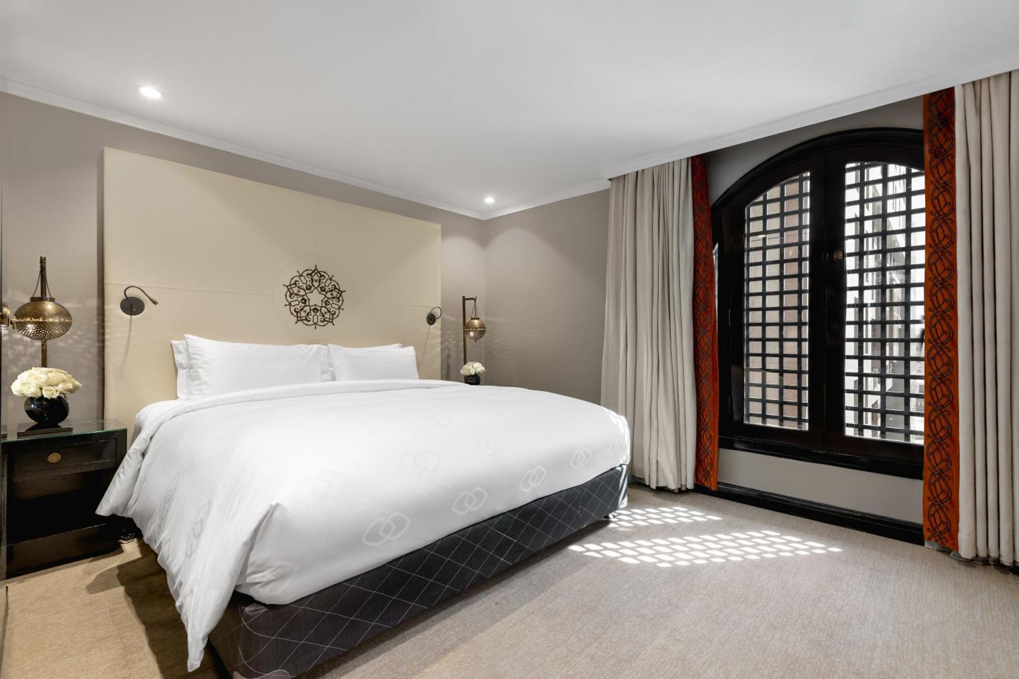 Sofitel Shahd Al Madinah Hotel Medina Buitenkant foto The photo shows a stylish hotel room featuring a large bed with white bedding positioned against a light-colored wall. Decorative elements include a circular wall piece above the bed and bedside lamps. The room has large windows with a unique grid pa