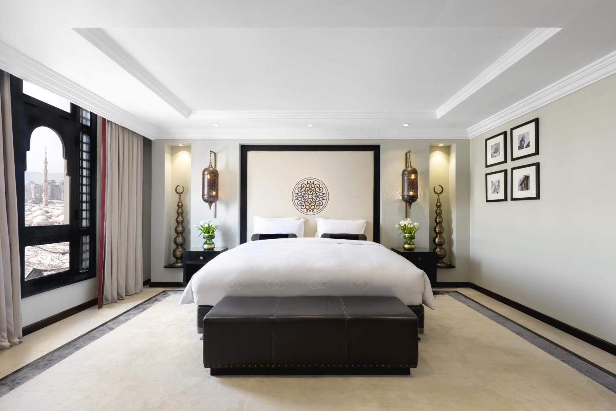 Sofitel Shahd Al Madinah Hotel Medina Buitenkant foto The photo shows a modern and stylish bedroom. It features a large bed with white linens, placed against a decorative wall with a circular design. On either side of the bed, there are elegant wall lamps illuminating the space. The room has a soft colo